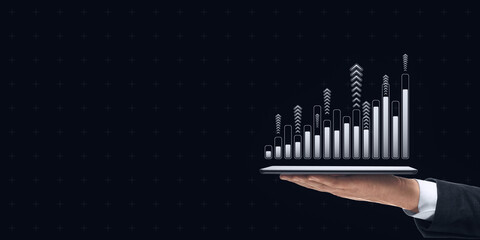 Hand holding tablet displaying upward bar chart with arrows on dark background, symbolizing financial growth and technology.