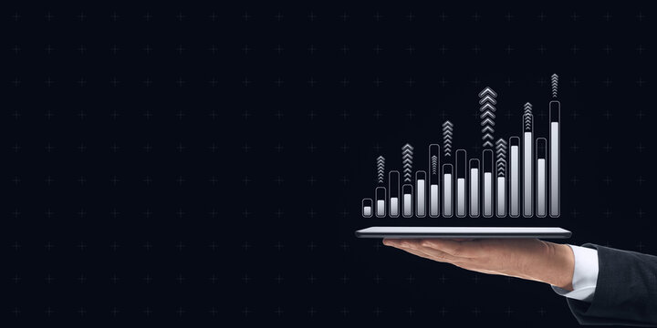 Hand holding tablet displaying upward bar chart with arrows on dark background, symbolizing financial growth and technology.