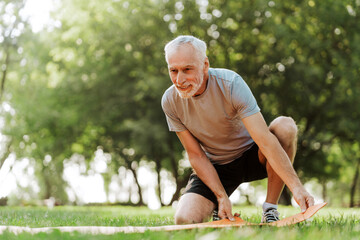 Naklejka premium Happy senior man unrolling yoga mat in the park getting ready for outdoor workout
