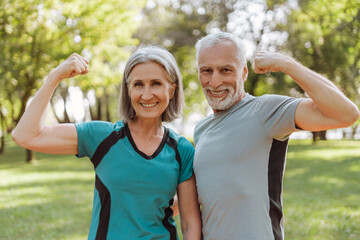 Fototapeta premium Smiling, attractive senior athletic couple flexing muscles in park, looking at camera outdoors