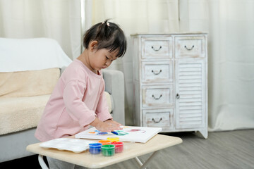 Naklejka premium Asian toddler girl deeply focused on finger painting with vibrant colors. Standing at a table, exploring artistic expression. Early childhood education with paint pots and canvas.