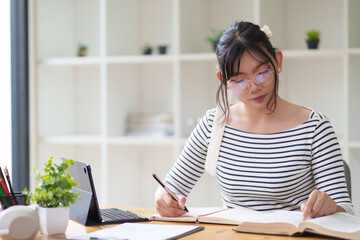 A young woman wearing glasses and a striped shirt is sitting at a desk, writing in a notebook. She appears focused, with an organized indoor setting around her and shelves in the background