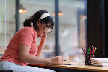 A young woman in a pink shirt is sitting by a window, writing in a notebook. She appears focused, with headphones around her neck and soft natural light illuminating the indoor space