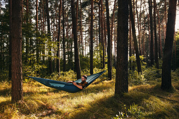 Wide shot of relaxed man using phone in a hammock in forest during autumn hike