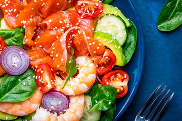Healthy food salmon salad for keto diet with shrimp, avocado, spinach, cucumber, tomato, cashew, sesame seeds. Blue background, top view