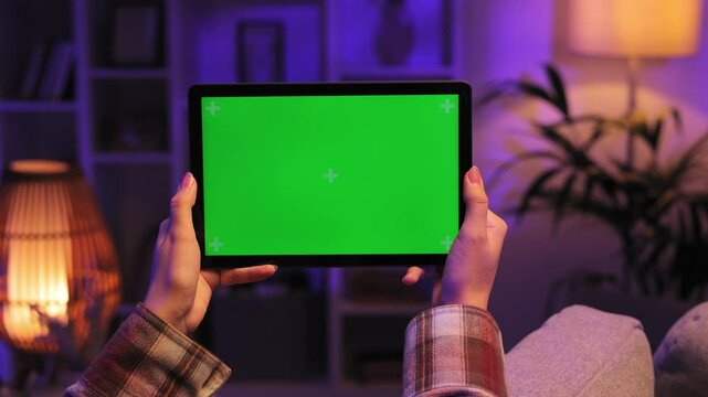 Closeup shot of girl lying on sofa using tablet in horizontal position with green screen mockup in evening room using different gestures while watching video.