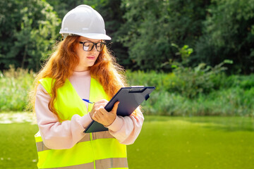 Female ecologist in overalls near a forest lake notes the ecological situation in the environment....