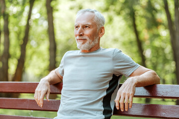 Happy senior man relaxing on bench after exercising outdoors looking away