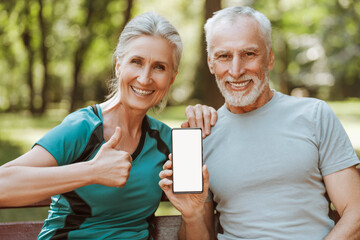 Happy senior couple showing smartphone with mockup screen sitting on bench in park, copy space