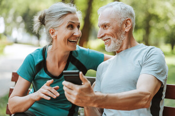 Happy senior couple using smartphone sitting on bench in park, ordering, buying, social media