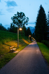 A nighttime view of a sidewalk and cycling path illuminated by street lamps. A bench sits quietly at the edge, while the distant Dolomites are faintly visible under the night sky