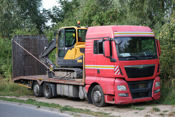 Red tow truck with excavator at the back is standing on street.