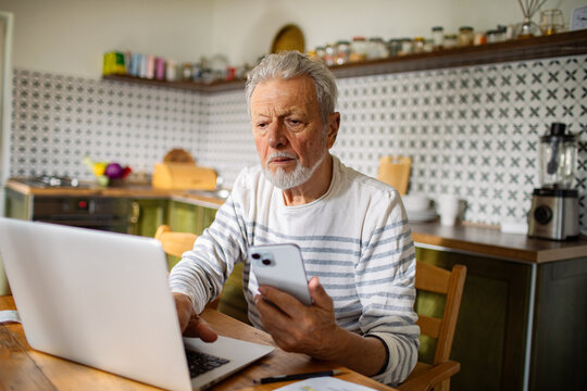 Elderly man managing bills and finances on laptop in kitchen