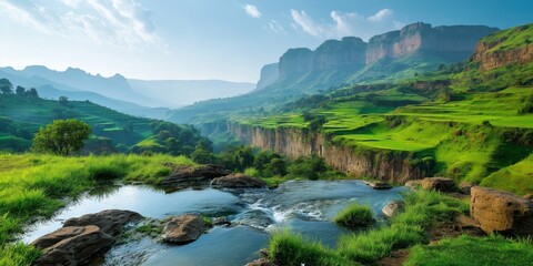 A beautiful mountain landscape with a river running through it. The water is calm and the grass is lush and green. The sky is clear and the sun is shining brightly. The scene is peaceful and serene