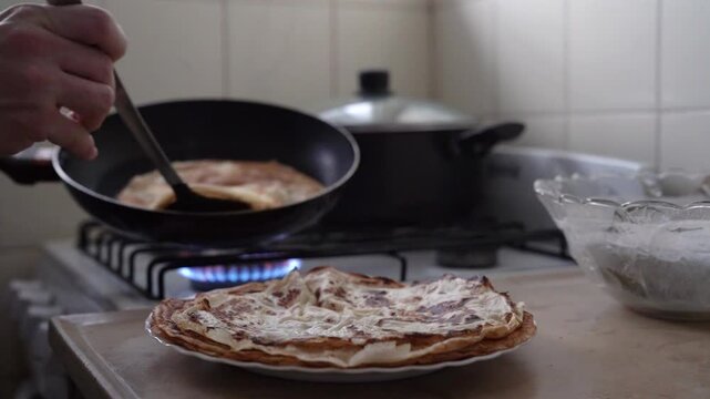 Making pancakes at home. Closeup view fo a male hand removing a fresh crepe heated in a pan, with a spatula, in the kitchen