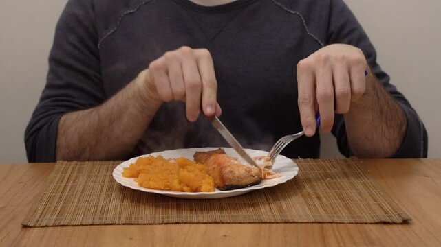 Dining fish. Closeup view of a caucasian man holding a knife and fork, cutting and eating a pink salmon filet with pumpkin puree
