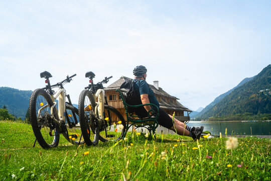 &Auml;ltere, dickerer Mann macht Pause auf einer Bank mit seinen Fahrr&auml;dern dahinter ein Holzhaus am Ossiacher See in Villach in K&auml;rnten &Ouml;sterreich