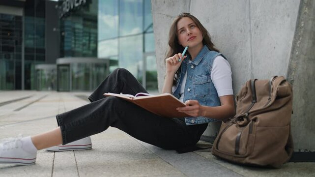 Video of focused beautiful student in front of university building or campus, studying outdoors, reading textbook, preparing for final exam.