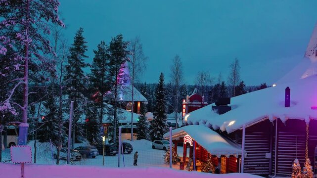 Nestled in a winter wonderland, charming cabins glow under colorful lights as dusk settles over a peaceful snowy village surrounded by tall evergreen trees.	