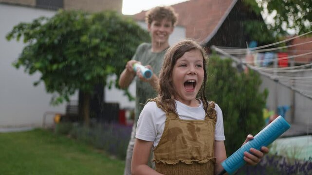 Siblings playing with water guns, water shooters during hot summer day. Children spending summer break outdoors on backyard.