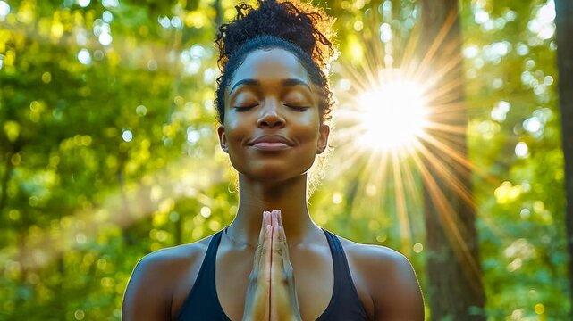 A young afro-american woman meditates with hands in prayer pose in a serene forest. Sunlight filters through the trees, creating a peaceful ambiance. She's wearing a dark tank top, and it's likely