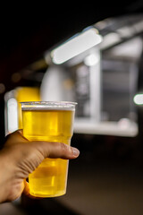 Close-up of a hand holding a plastic cup filled with beer, with a blurred background of a food truck at night. Perfect for themes of street food, nightlife, and casual dining.
