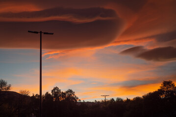 Paisaje. Nube densa al atardecer, con un poste de luz con arboles al fondo.	
