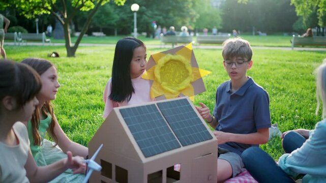 Children learning about renewable energy and solar panels during sustainable education class outdoors, using cardboard model of house wit solar panel on roof.