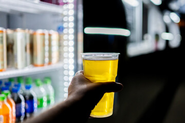 A hand holding a clear plastic cup filled with a yellow beer, set against a blurred background of colorful bottled drinks in a refrigerated display. Ideal for themes of refreshment, beverages