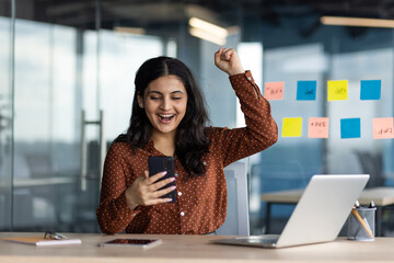Joyful successful business woman at workplace holding phone in hands received notification of...