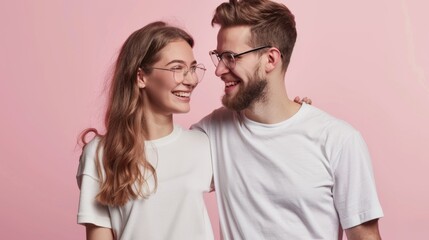 Fototapeta premium A smiling young couple in white t-shirts stand against a pink background, radiating joy and affection as they look at each other.