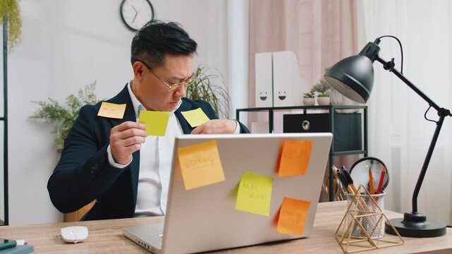 Tired exhausted Asian businessman working on laptop at office with many sticker task inscriptions. Freelancer man busy occupation feels sad boredom overworked multitasking. Panic attack, deadline
