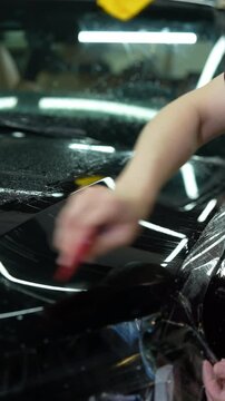 Closeup vertical shot of auto detailer meticulously applying protective vinyl film to luxury car hood in professional detailing workshop, ensuring precision and care for flawless finish, slow motion.