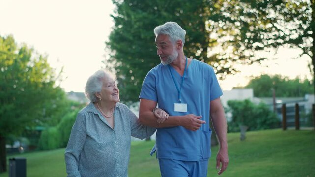 Male caregiver with elderly patient in park. Geriatric nurse accompany lovely elderly woman on outdoor walk, enjoying conversation.