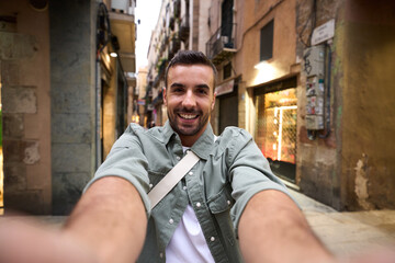 POV Young handsome man smiles taking selfie on Barcelona city touristic street in background. Millennial male tourist person takes happy photo of himself holding his phone with both hands outdoors