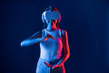 Female standing surrounded by neon light wear VR headset connecting metaverse, futuristic cyberspace community technology. Elegant woman using both hand interacting with virtual object. Hallucination.