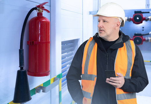 Enterprise safety engineer. Man near fire extinguisher. Inspector checks presence fire extinguisher. Man industrialist holding phone. Guy employee industrial company. Safety engineer in orange vest