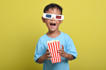 Fototapeta premium Excited Asian boy wearing 3d glasses, Watching movie while holding popcorn bucket isolated on yellow background. Concept of watching movie and cinema