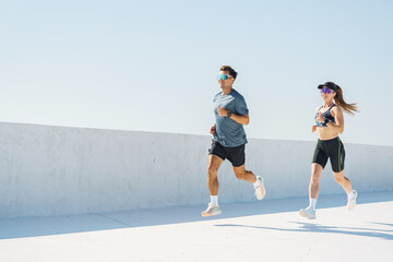 A man and woman are jogging in a minimalist outdoor space under bright sunlight, casting long...