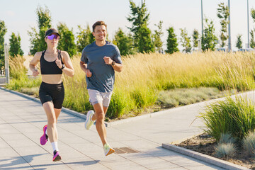A man and woman jog along a path surrounded by tall grass and trees on a sunny day, enjoying the...