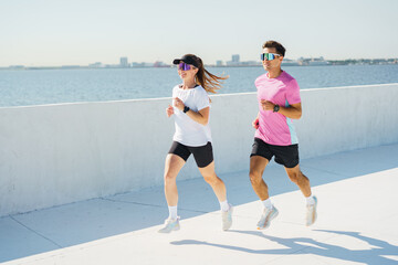 A man and woman jog along a sunny waterfront, both wearing bright athletic gear and sunglasses.