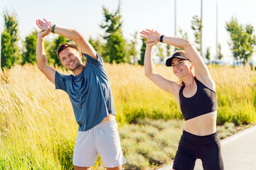 A man and woman stretch together outdoors in a sunny, grassy area, smiling and enjoying the warm...