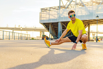 A man in bright athletic wear stretches outdoors at sunset, preparing for a workout, with modern structures and a bridge in the background.