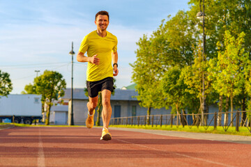 Obraz premium A man in a yellow shirt runs on a sunlit track surrounded by greenery, with a determined smile.