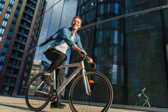 A man rides his bicycle through an urban business district, smiling confidently in the sunlight.