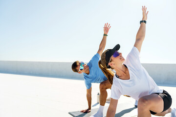 A man and a woman stretch outdoors, reaching upward during a sunlit exercise routine.