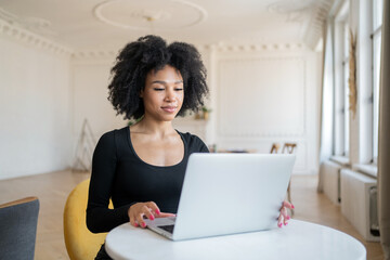 A focused woman with natural curls works on her laptop in a bright, elegant room, exuding calm.