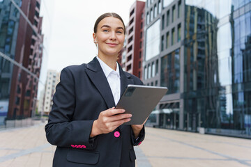 A confident businesswoman stands amidst modern glass buildings, holding a tablet, with a smile.