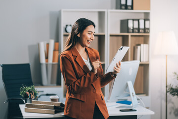 Attractive smiling Asian businesswoman standing holding tablet working and recording work details in office.