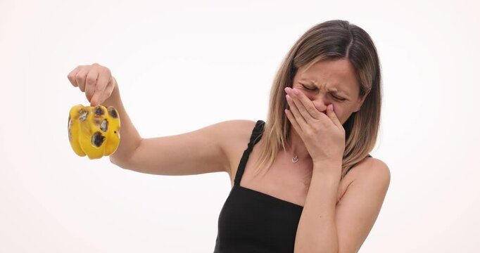 Woman holds rotten bell pepper covering nose with hand on white background. Blonde female experiences disgust with unpleasant smell of vegetable